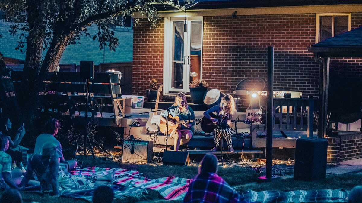 Two performers sit on an outdoor porch, playing music for a large crowd